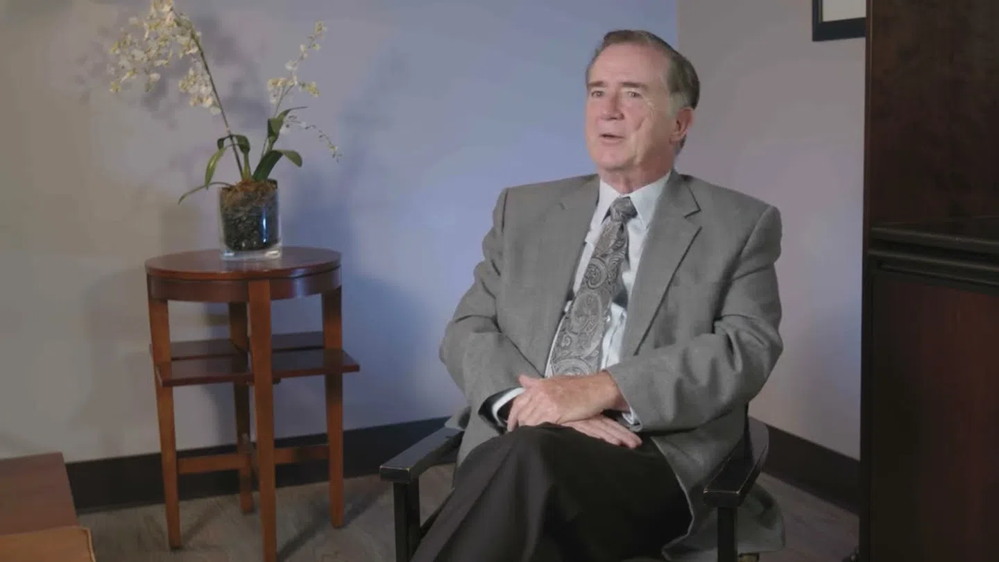 Man in gray suit sitting on chair in an office setting with a wooden table and white orchid plant.