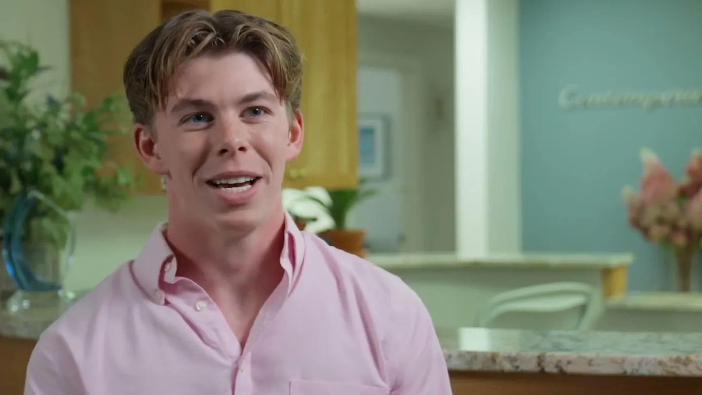 Smiling young man with light brown hair wearing a pink shirt indoors, with plants and kitchen background.