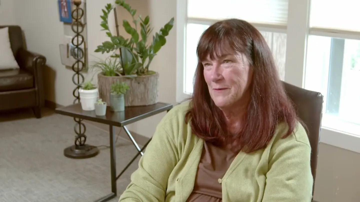 Middle-aged woman with brown hair in green cardigan smiling indoors with plants and chair in the background