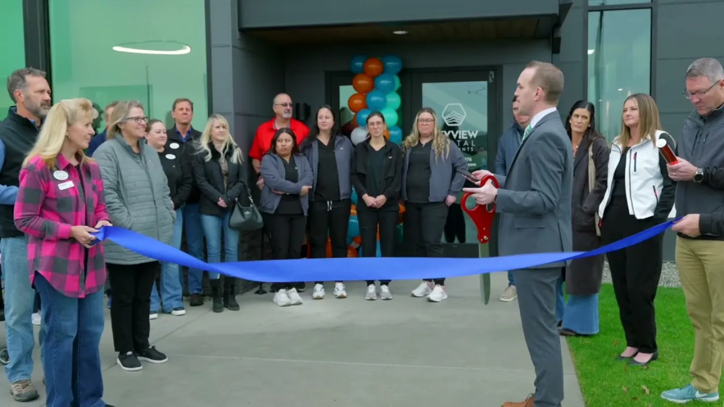 Group of people at a ribbon-cutting ceremony outside a dental office with balloons and large scissors.