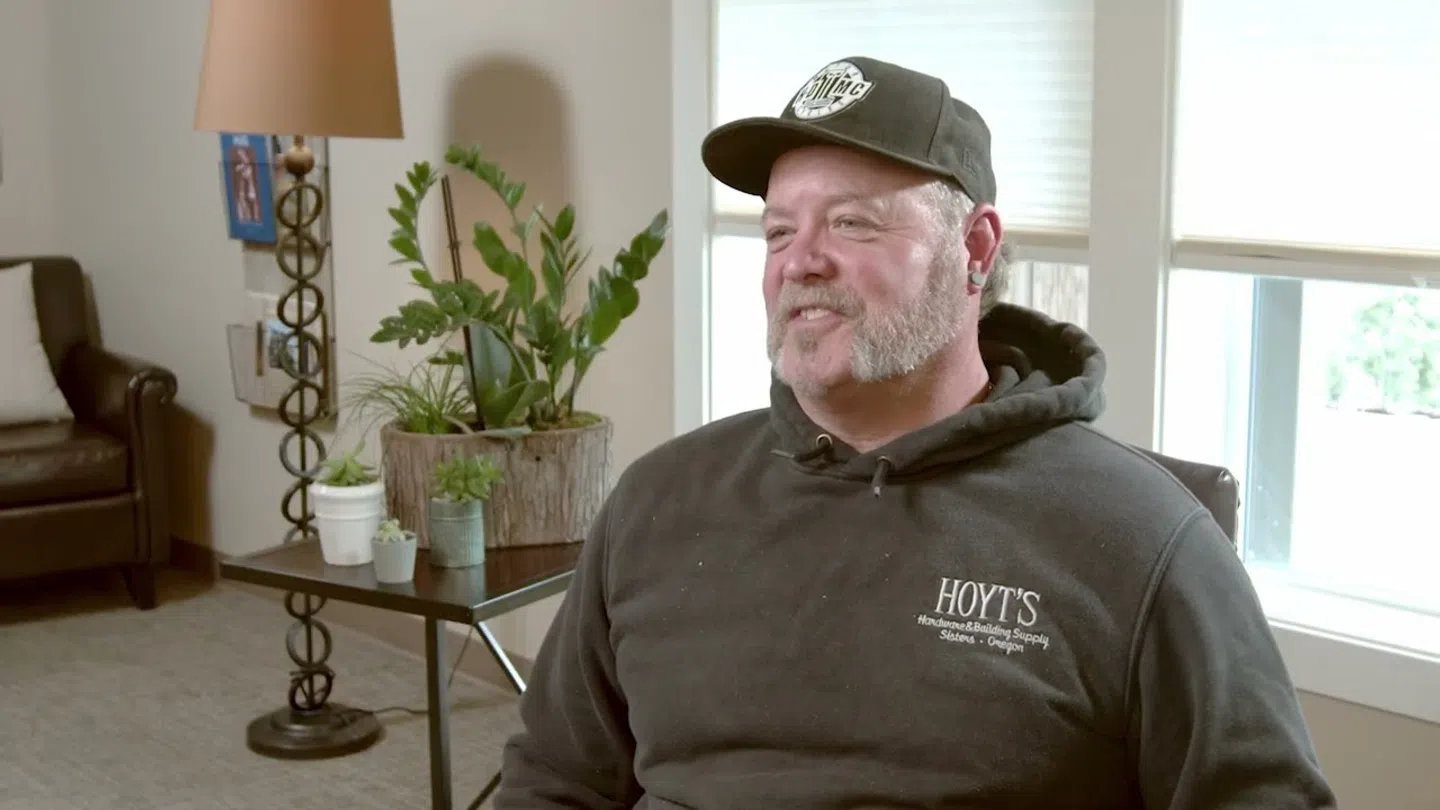 Man wearing a black Hoyt's hoodie and cap sitting in a room with plants and sunlight through windows