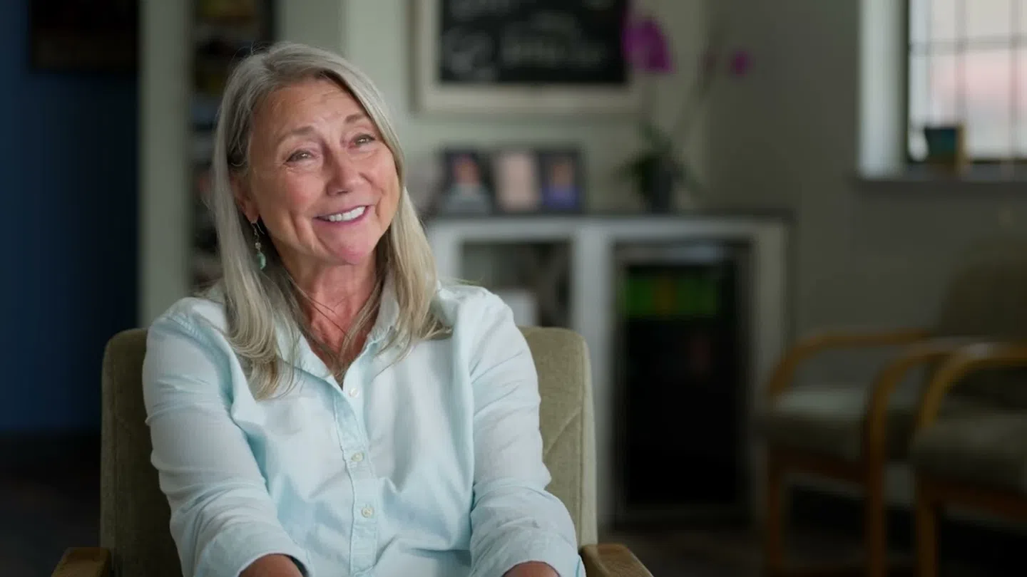 Smiling middle-aged woman with gray hair sitting in a cozy room with shelves and chairs in the background