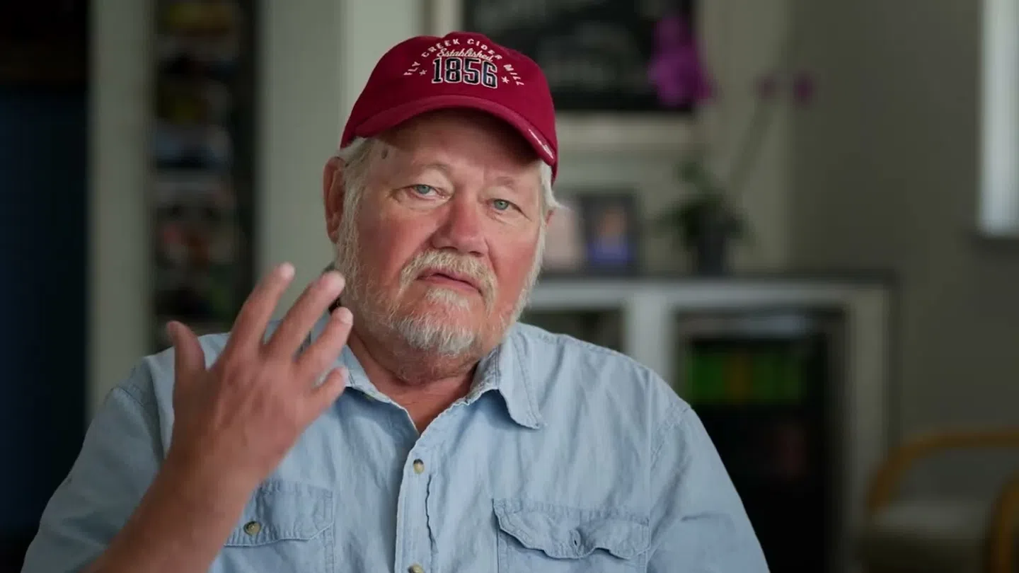 Older man with white beard wearing red cap and blue shirt gesturing with hand indoors.
