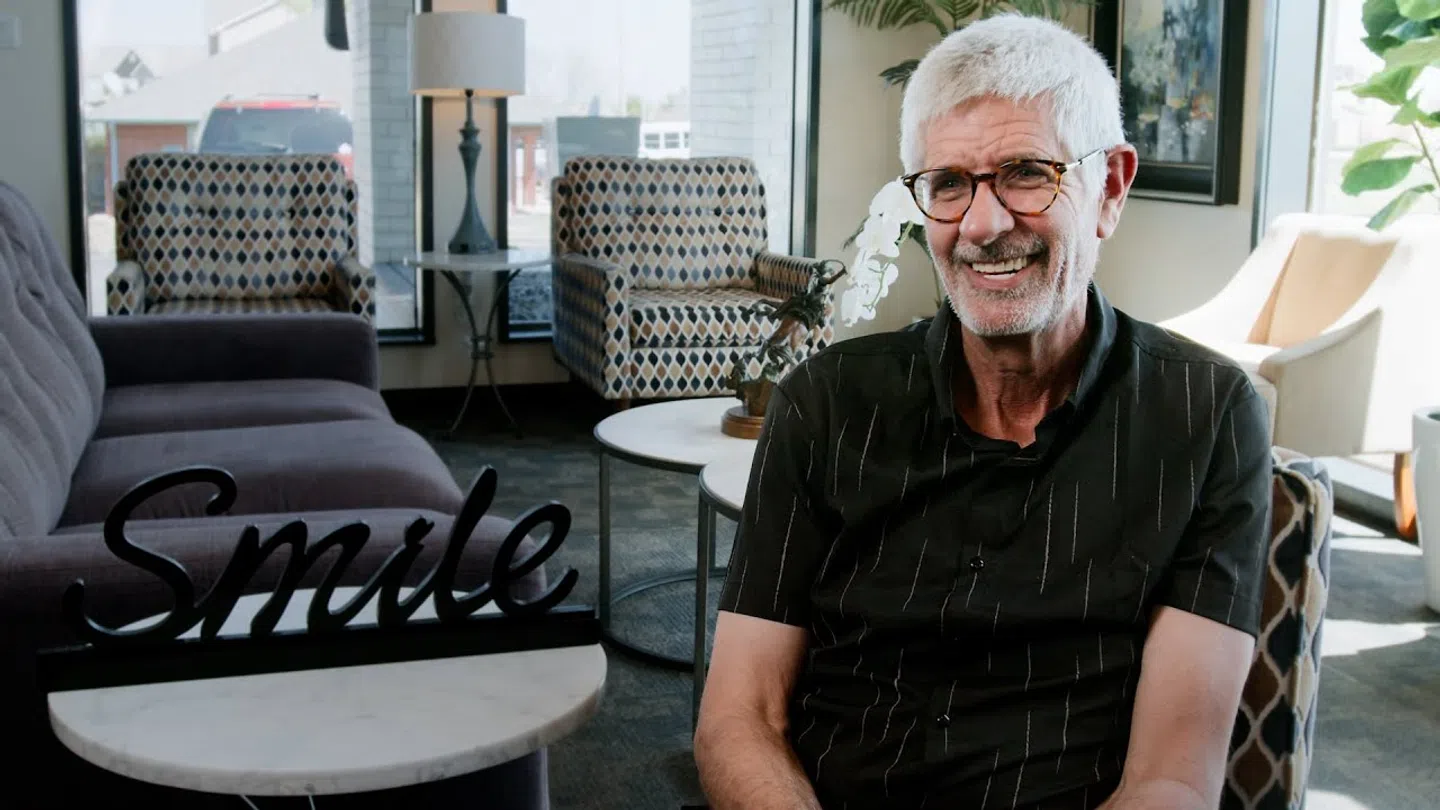 Smiling elderly man with glasses sitting in a modern living room with decor and a 'Smile' sign on a table.