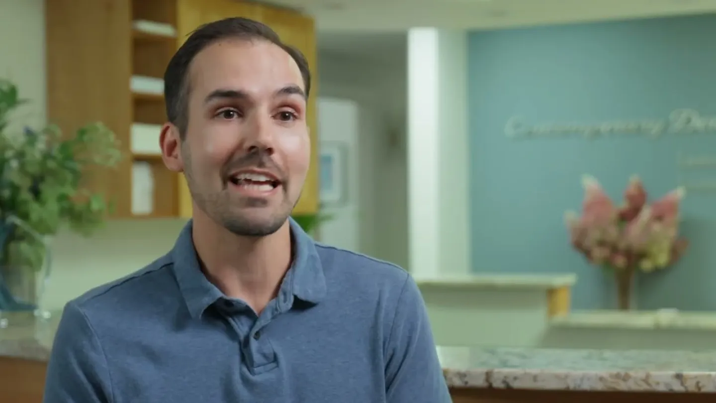 Man in blue polo shirt speaking inside a modern dental office with plants and flowers in the background