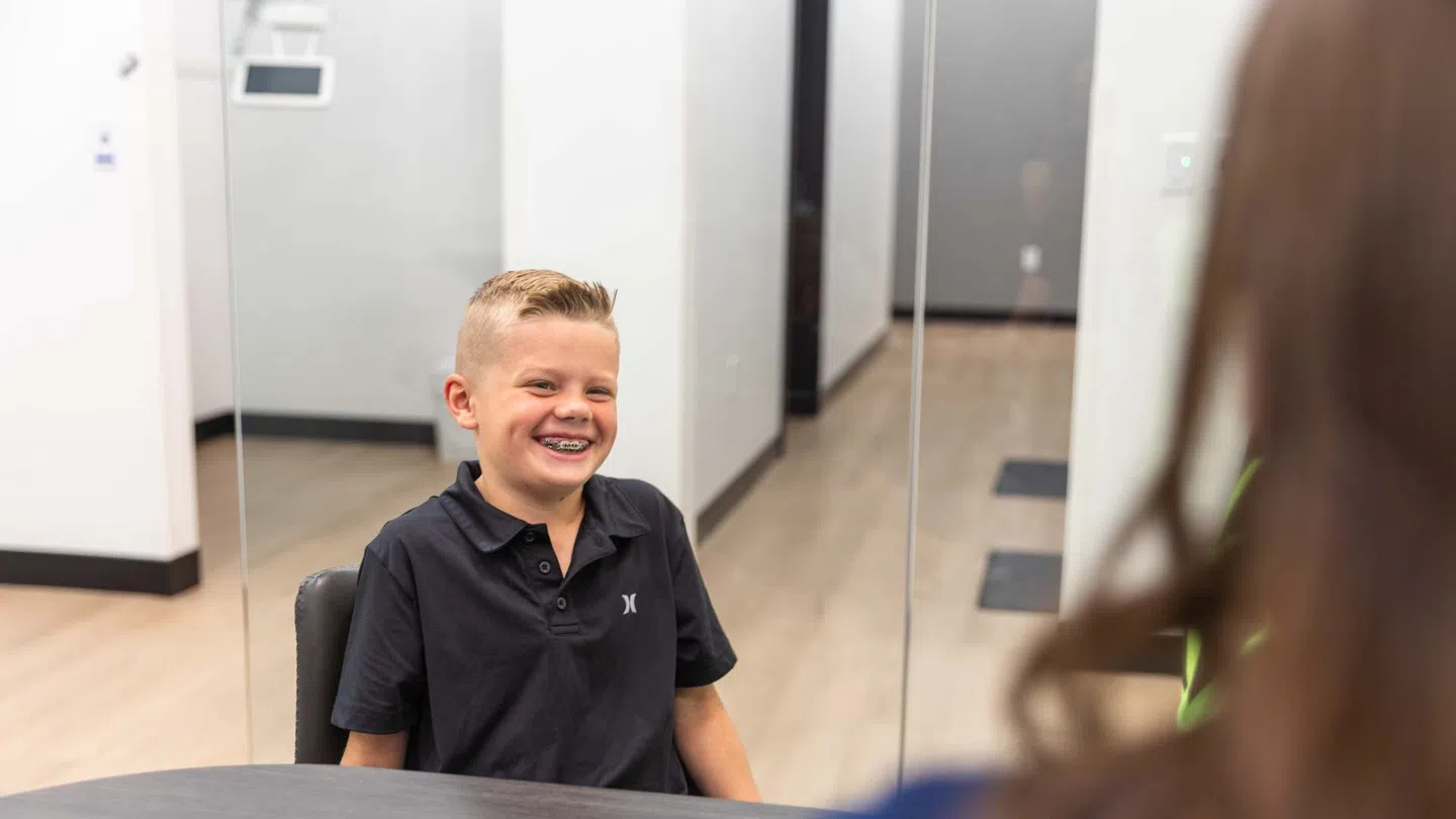 Smiling boy with braces sitting at a table in a modern office setting speaking with a person.