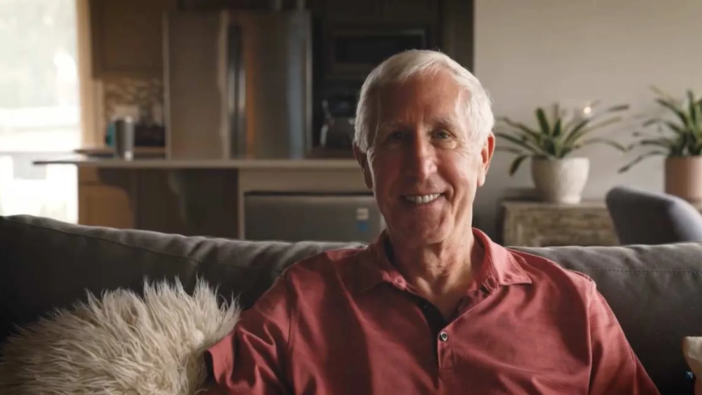 Smiling elderly man with white hair sitting on a couch in a cozy living room with plants and warm lighting.
