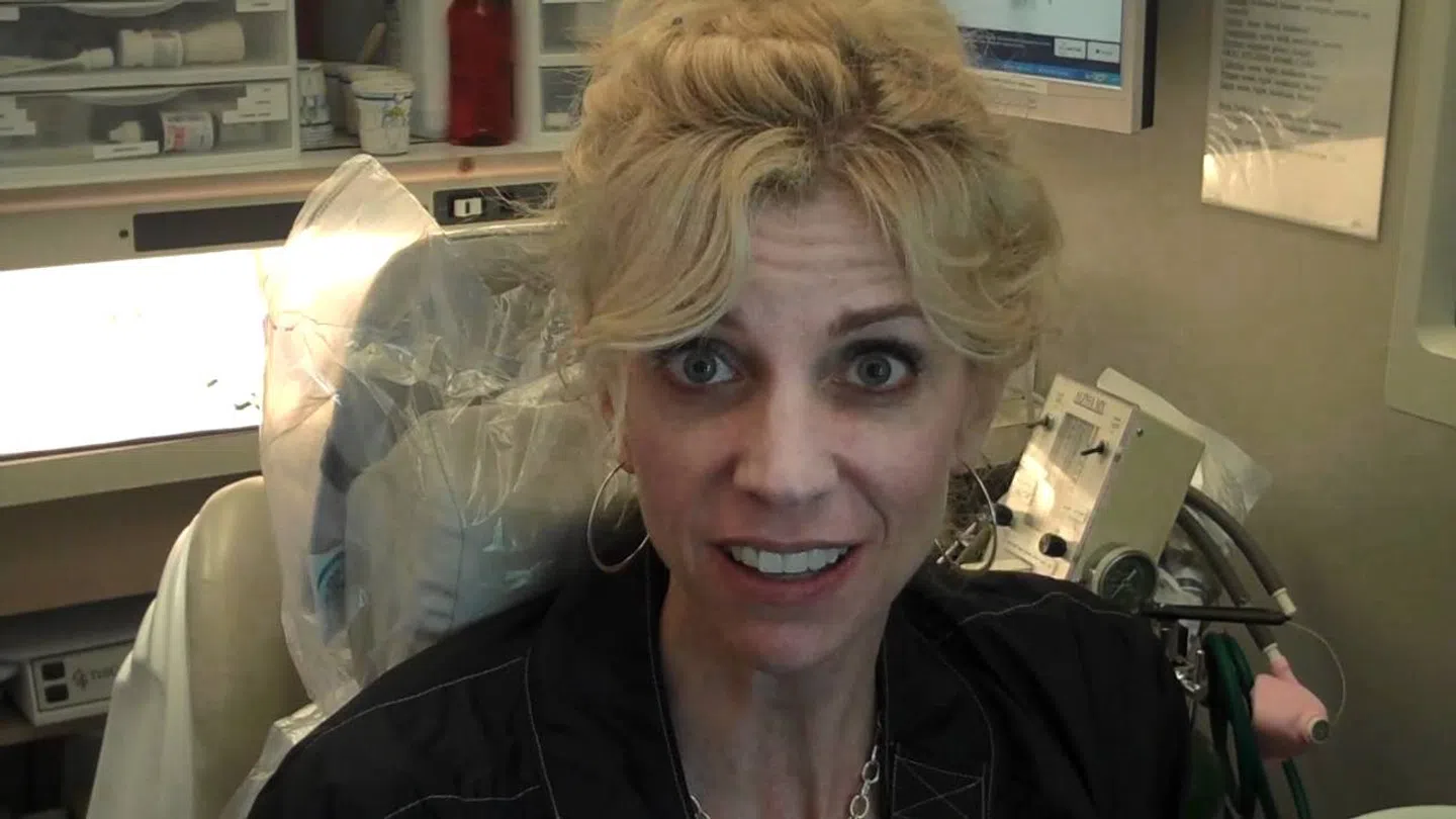 Smiling woman with blonde hair sitting in a medical or dental office surrounded by equipment and supplies.