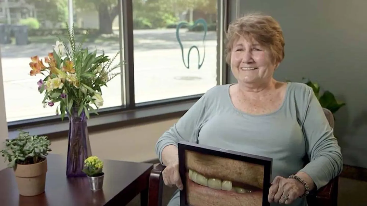 Elderly woman smiling and holding a photo of her teeth in a dental office with plants and tooth logo.