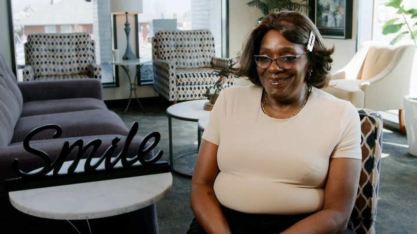 Smiling woman wearing glasses and a hair clip seated in a modern living room with a smile sign on the table.