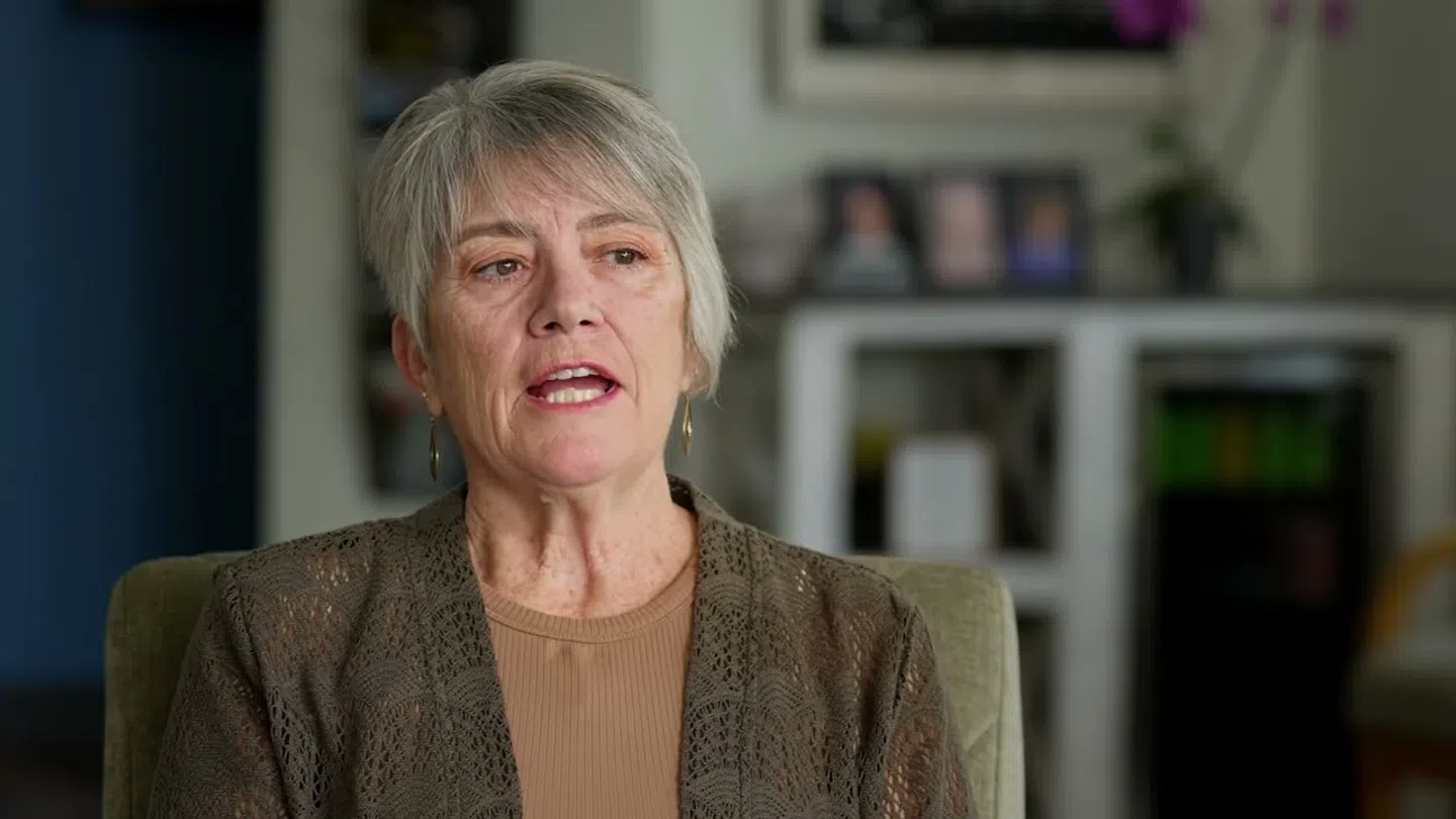 Middle-aged woman with short gray hair speaking indoors, wearing a brown top and lace cardigan in a cozy room.