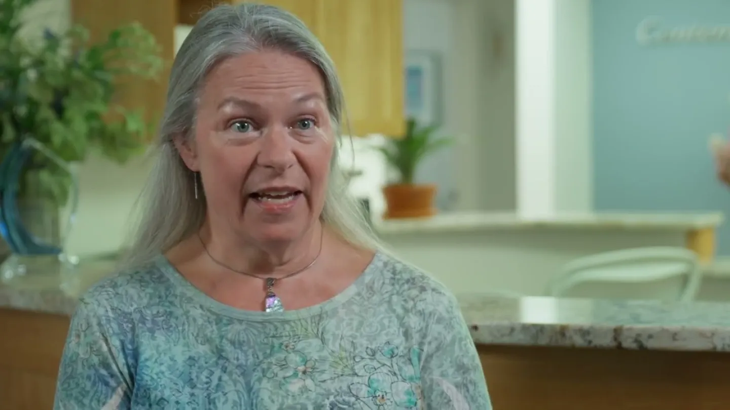 Middle-aged woman with gray hair wearing a patterned blue shirt speaks in a bright kitchen setting.
