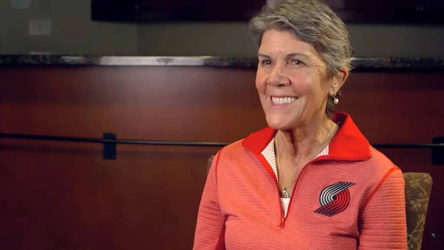 Smiling older woman wearing a Portland Trail Blazers pullover sitting in an indoor setting with a dark wood background.
