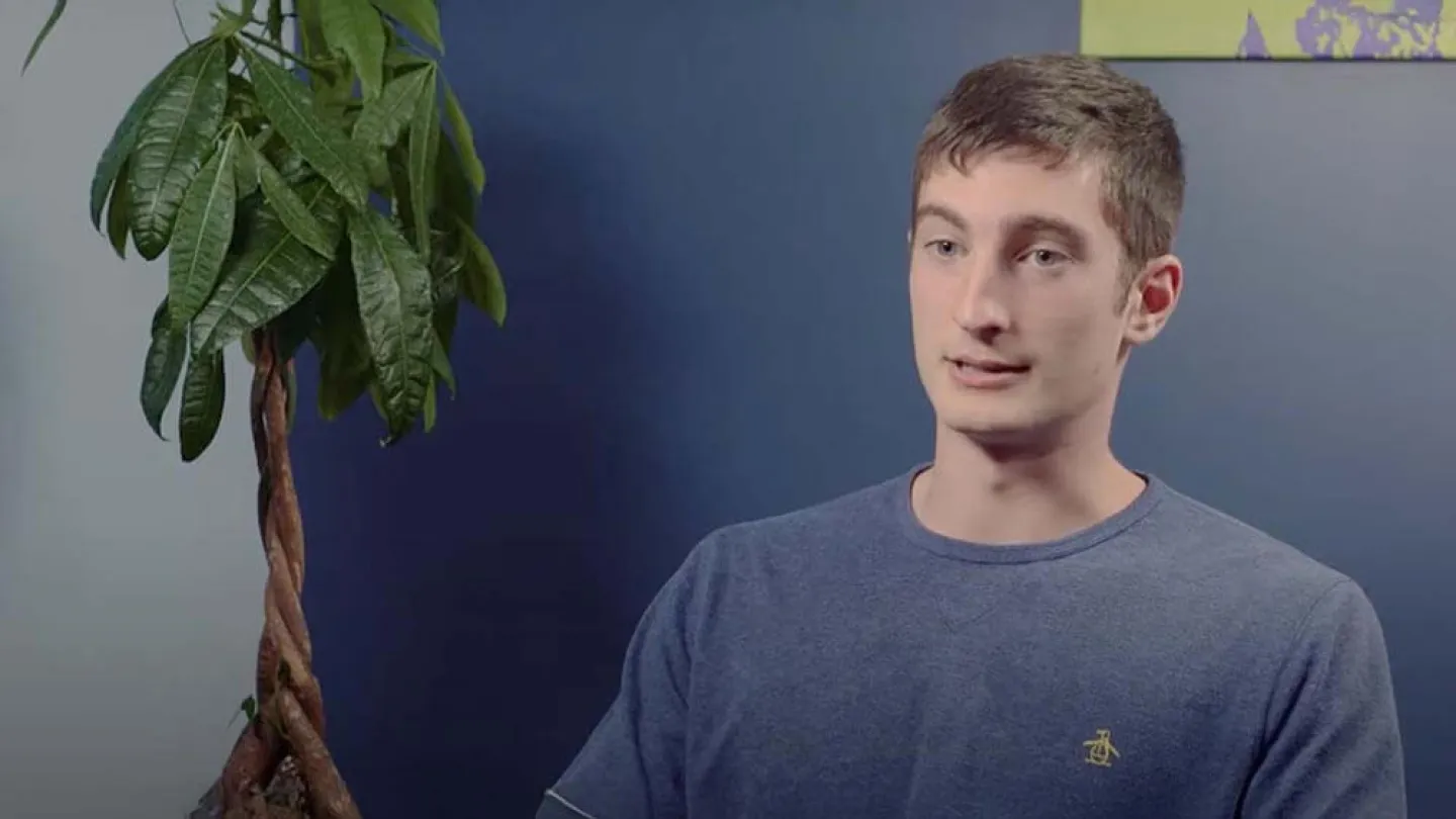 Young man in blue shirt seated indoors next to a braided money tree plant against a two-tone blue wall.