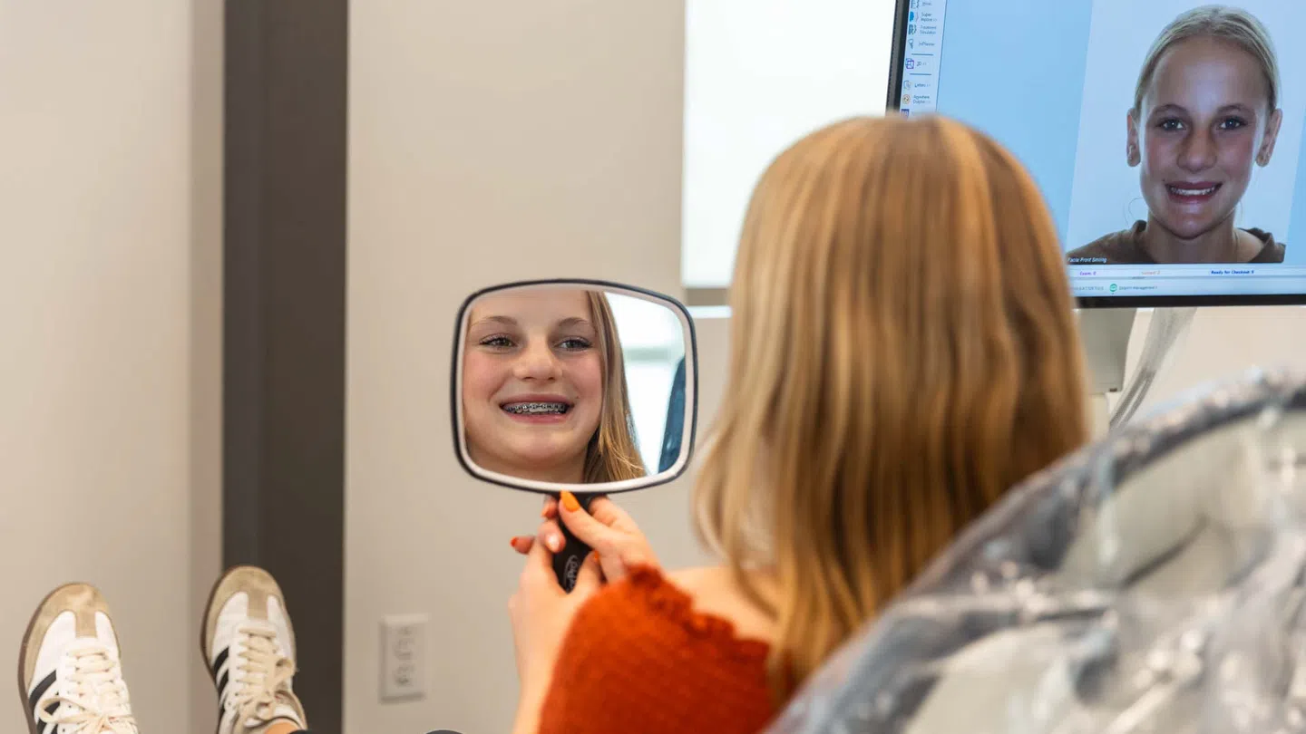 Girl with braces smiling while looking at her reflection in a handheld mirror at orthodontist office.