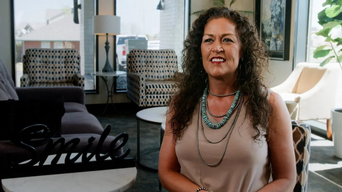 Woman with curly hair and layered necklaces smiling in a bright, stylish lounge area with modern furniture.