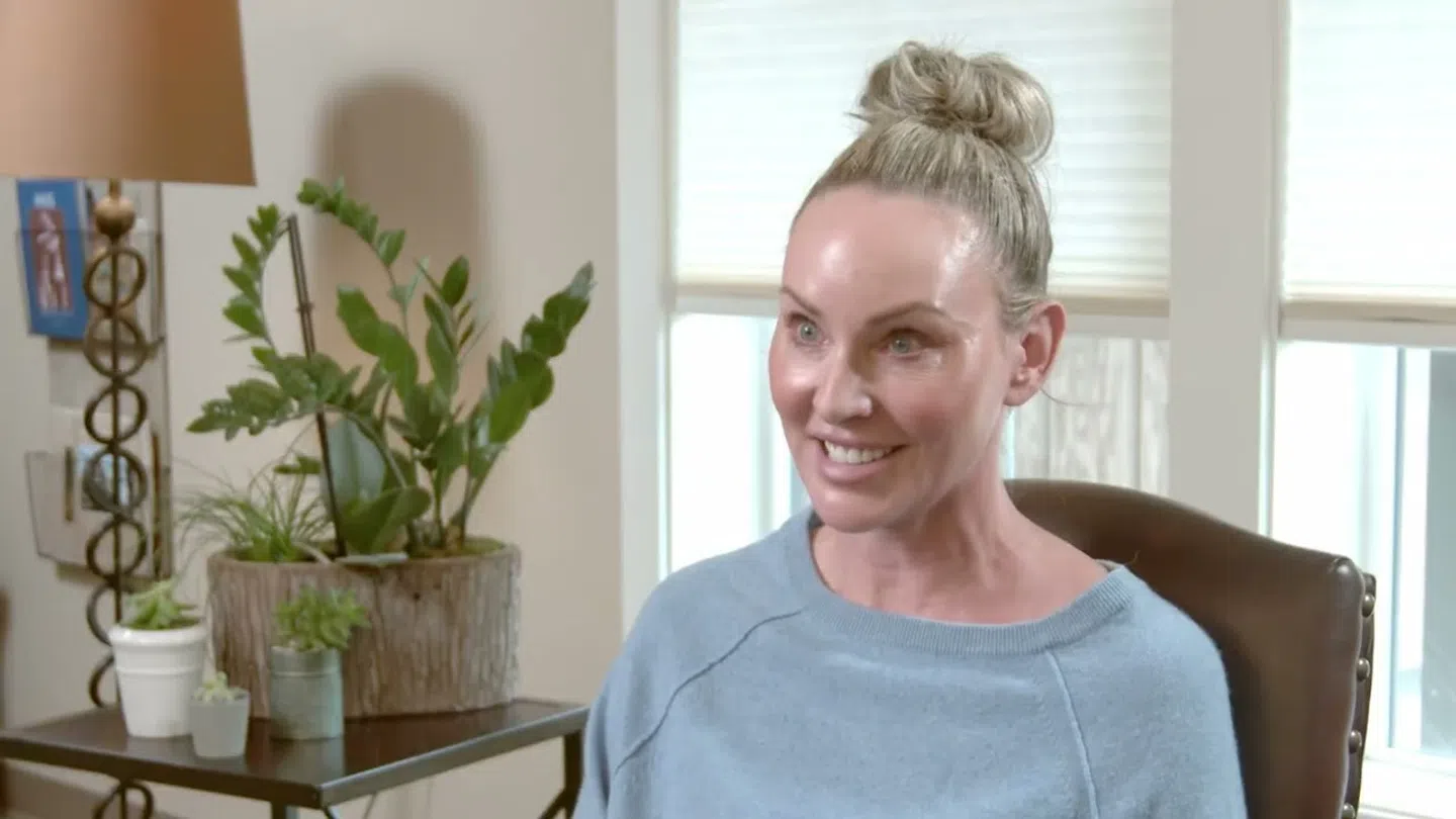 Smiling woman with blonde hair in a bun sitting in a chair near a plant and window blinds in a cozy room.