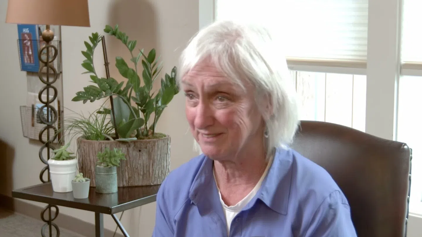 Smiling elderly woman with white hair wearing a blue shirt sitting in a cozy room with plants and natural light.