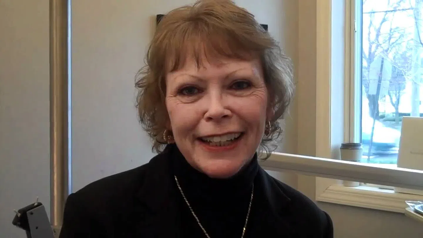 Smiling woman with short hair wearing a black top and necklace in a bright room with large windows.