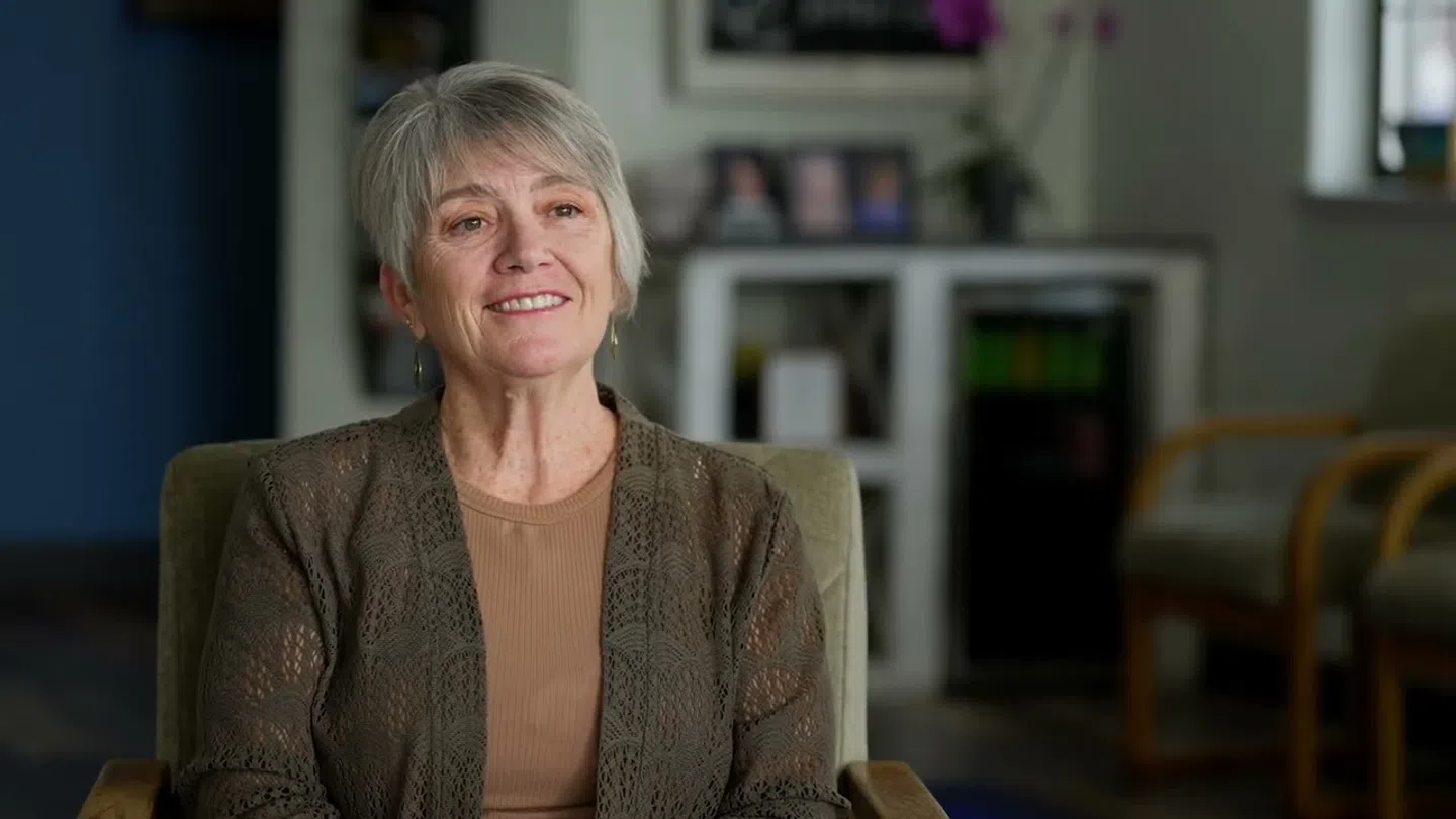 Smiling older woman with short gray hair wearing a brown top and cardigan sitting in a modern room.
