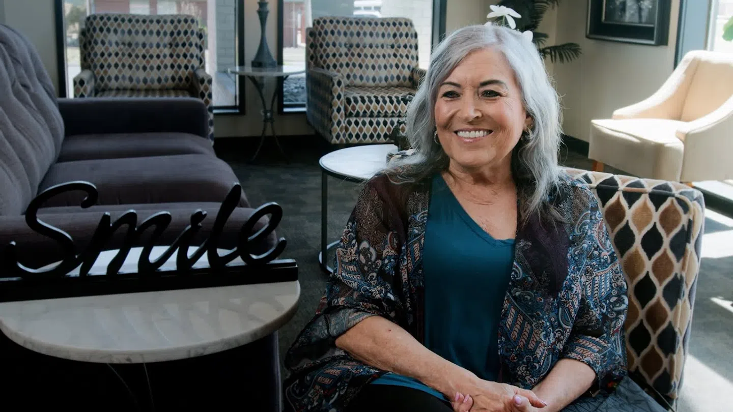 Smiling elderly woman sitting in a cozy modern living room with decor including a decorative Smile sign.