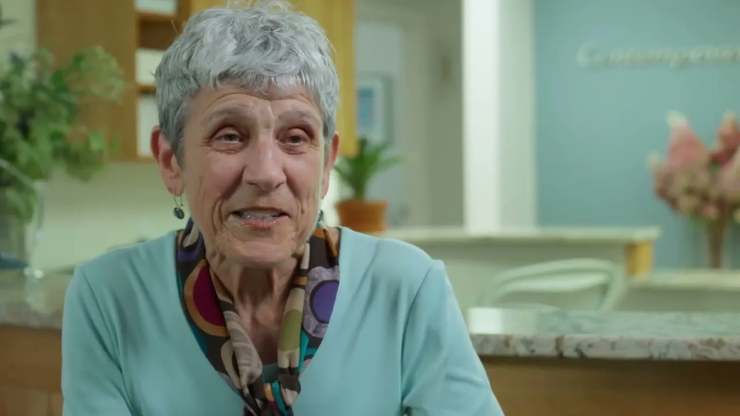 Elderly woman with gray hair and patterned scarf smiling indoors in a cozy, softly lit room.
