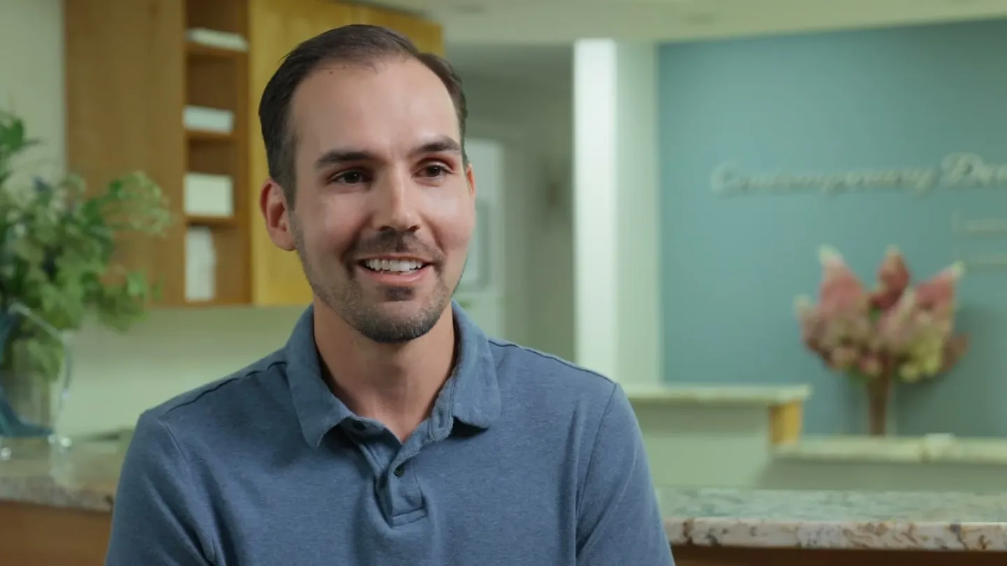 Man in a blue polo shirt smiling in a modern office with a plant and blurred background decor.