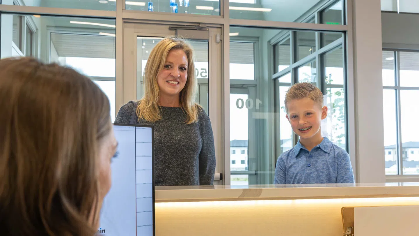 Woman and young boy smiling at reception desk inside modern building with glass doors and windows.