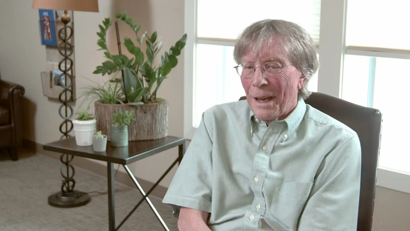 Elderly man with glasses sitting in a chair inside a well-lit living room with plants and a lamp.