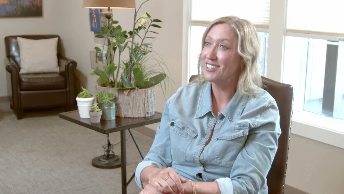 Smiling woman in denim shirt sitting in a cozy living room with plants and natural light from windows