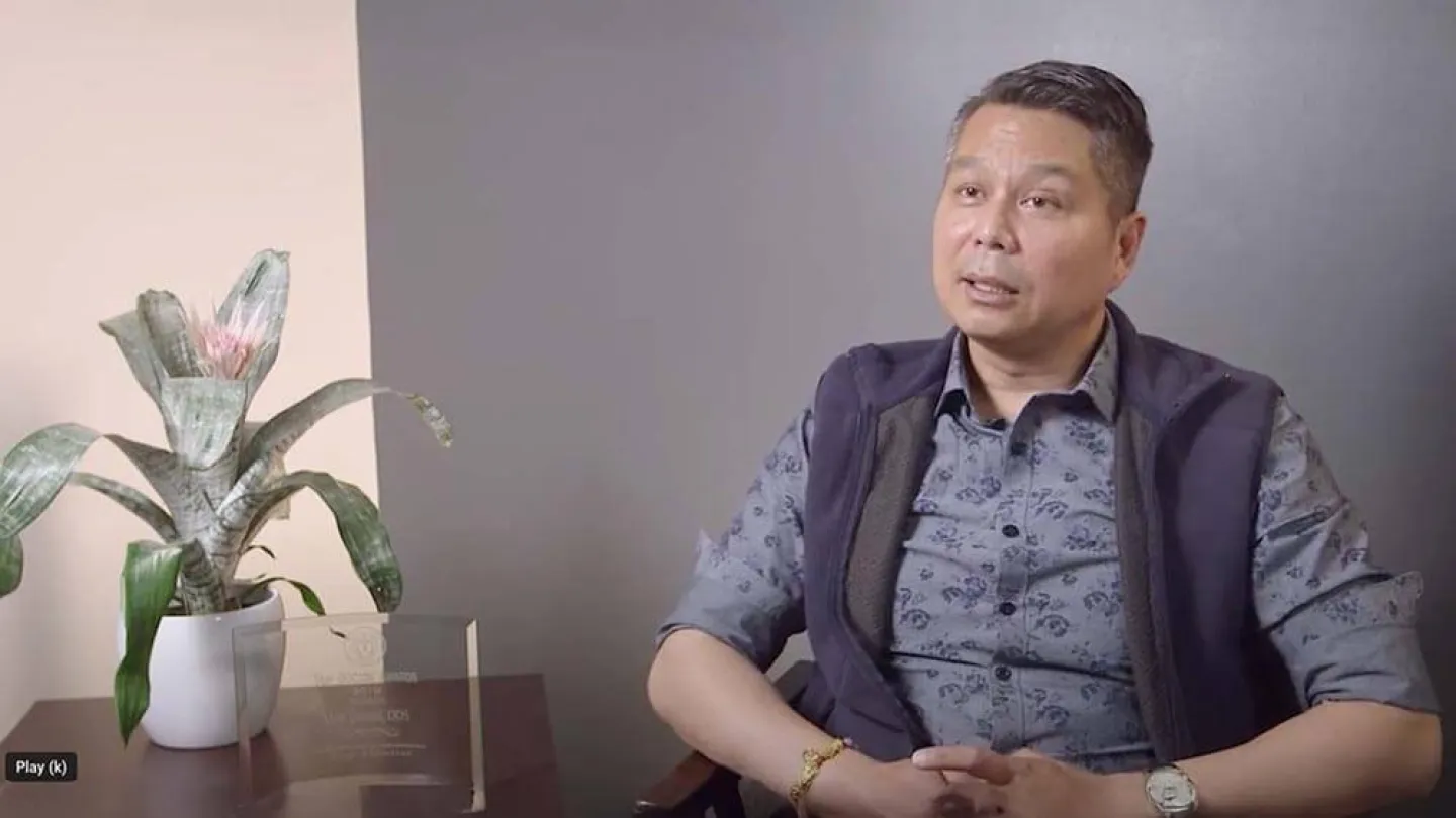 Man in patterned shirt and vest sitting in chair beside table with plant and award plaque in office.