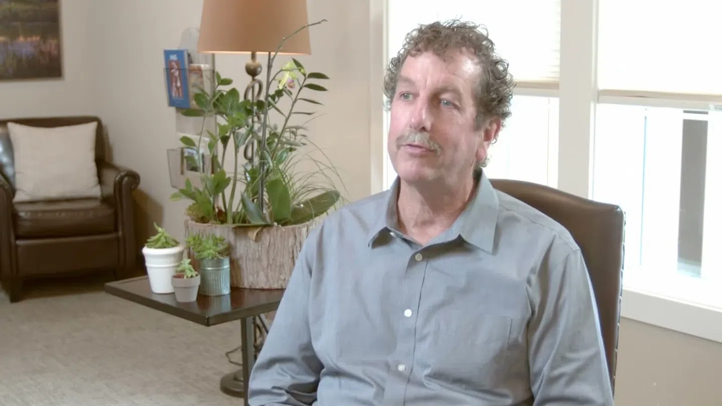 Man with curly hair and mustache sitting in a chair in a living room with plants and a lamp behind him.