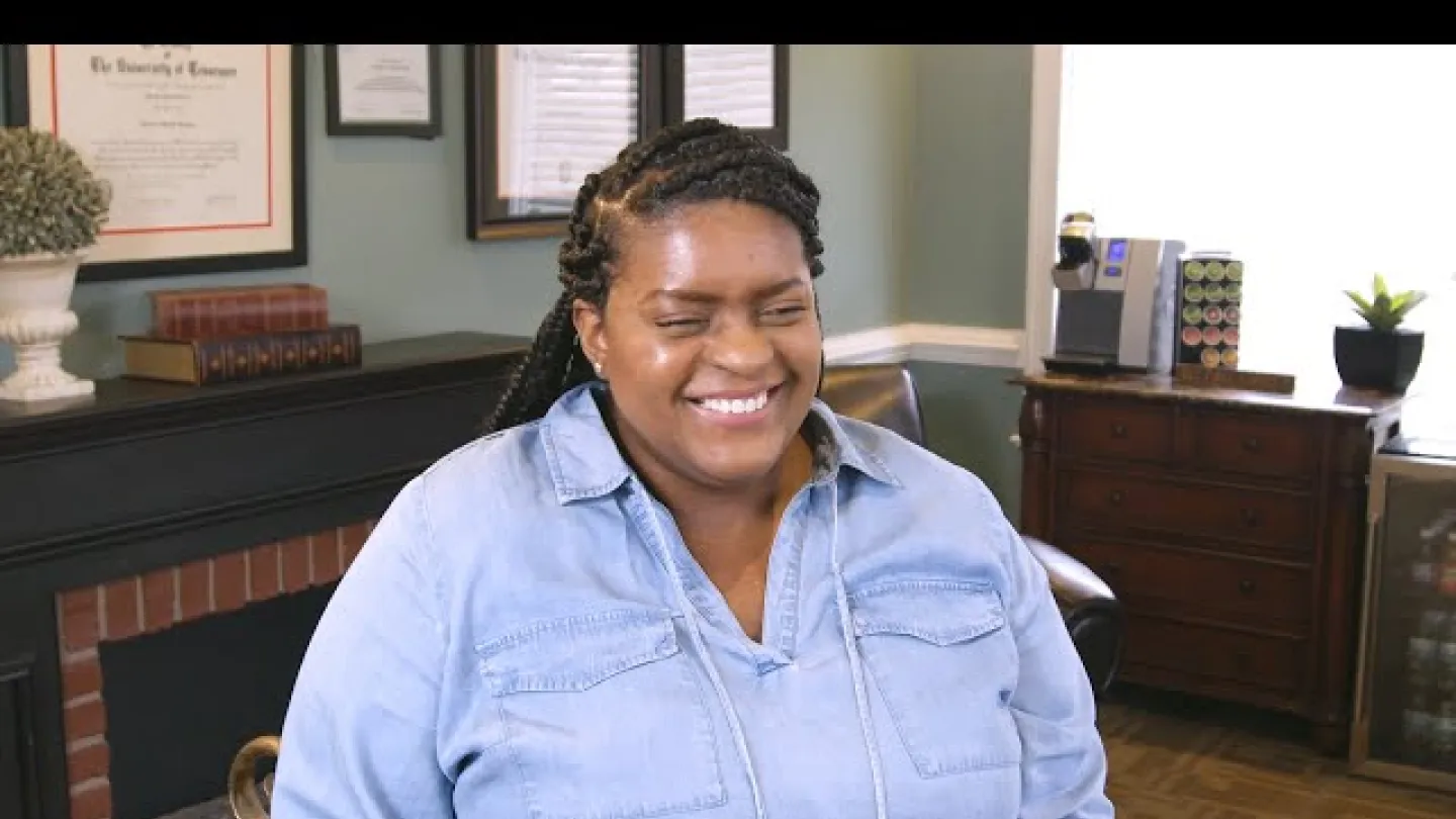 Smiling woman with braided hair in a denim shirt sitting in a cozy office with framed certificates and wooden furniture.