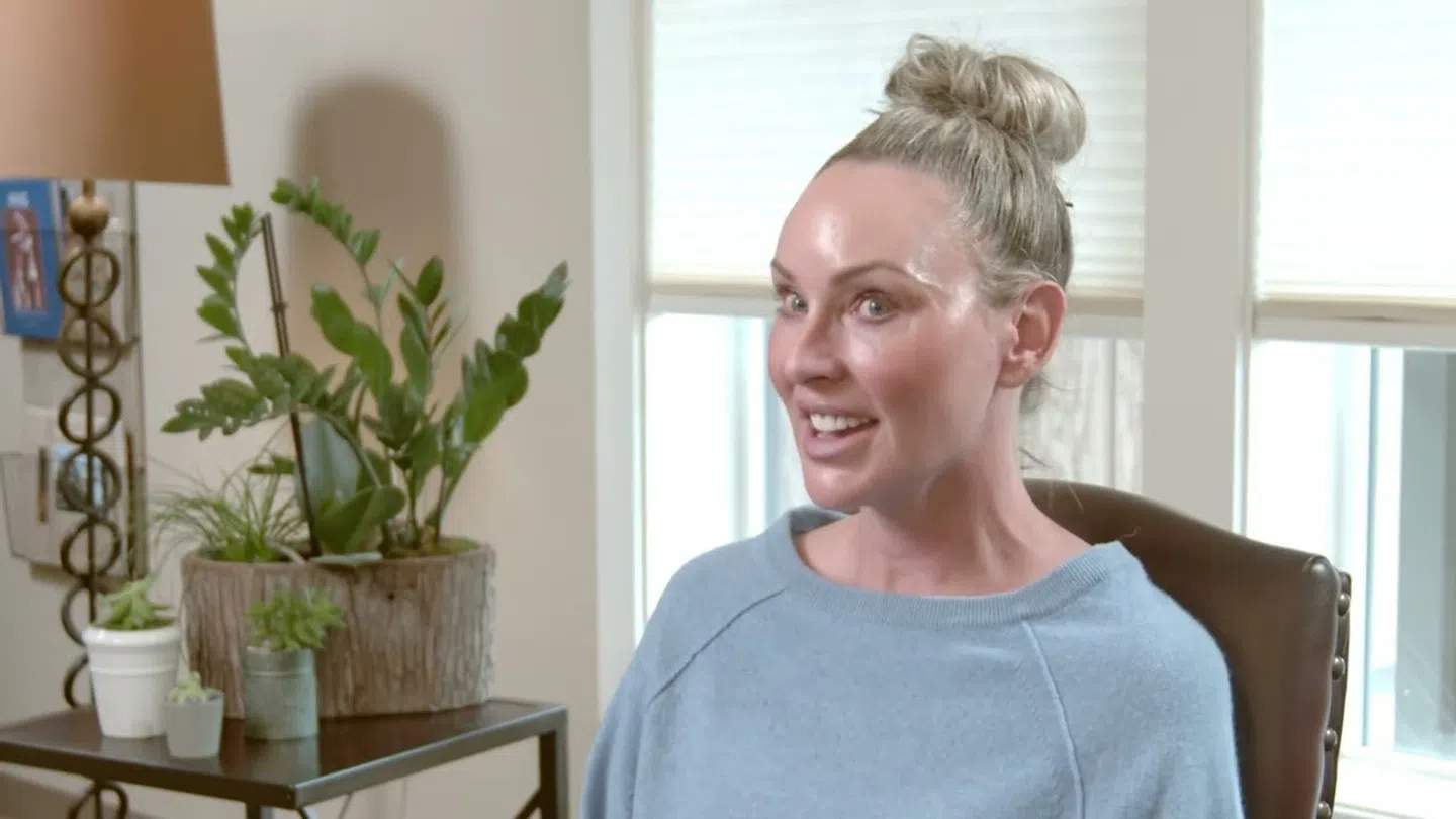 Smiling woman with blonde bun sitting in chair near window and indoor plants on side table.