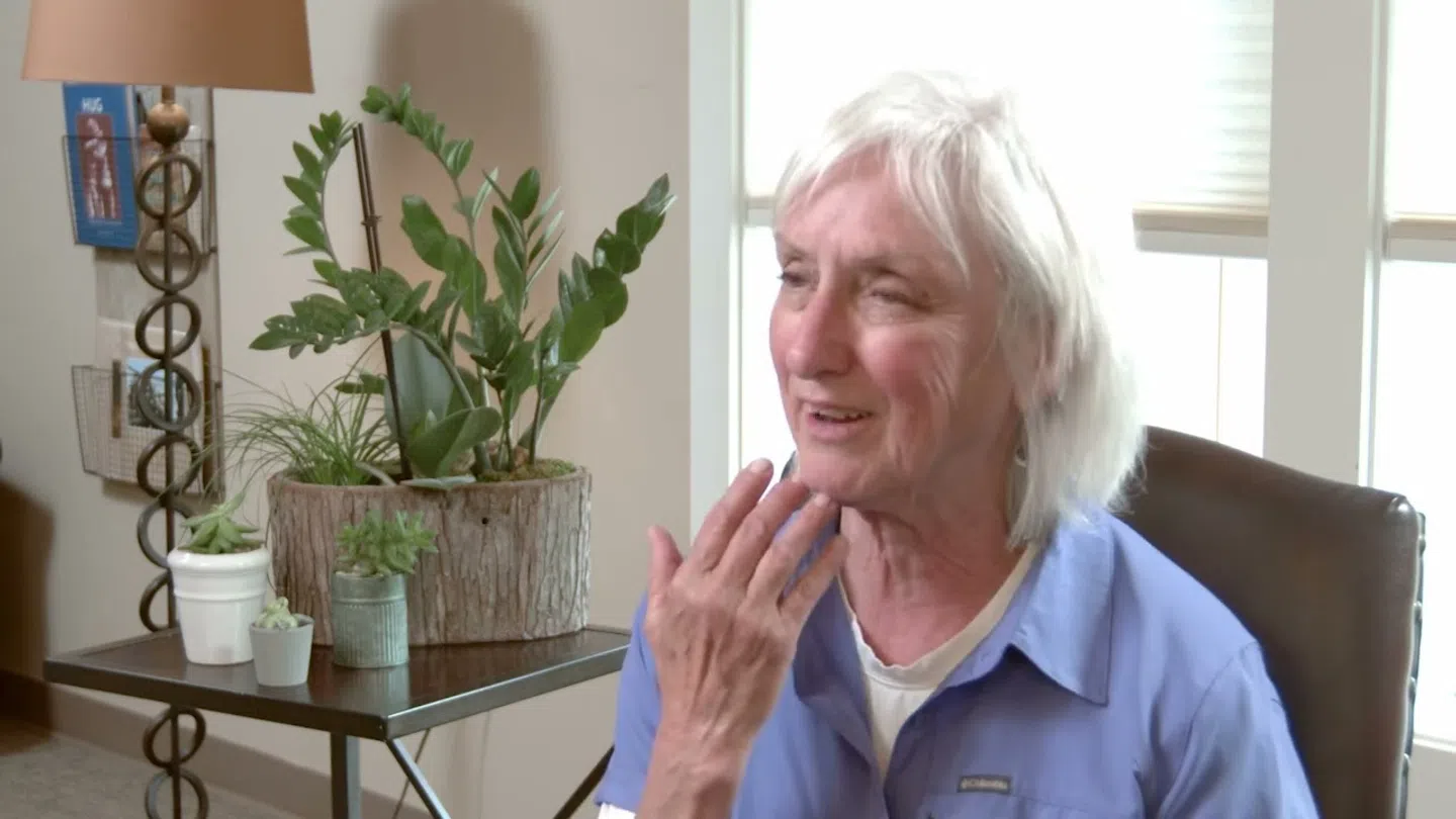 Elderly woman with white hair smiling and touching her face while sitting indoors near plants and a lamp.