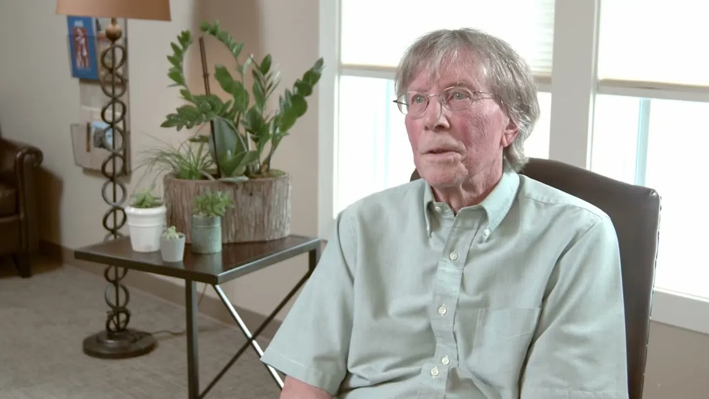 Elderly man with glasses sitting in chair near table with potted plants in bright room with large window.