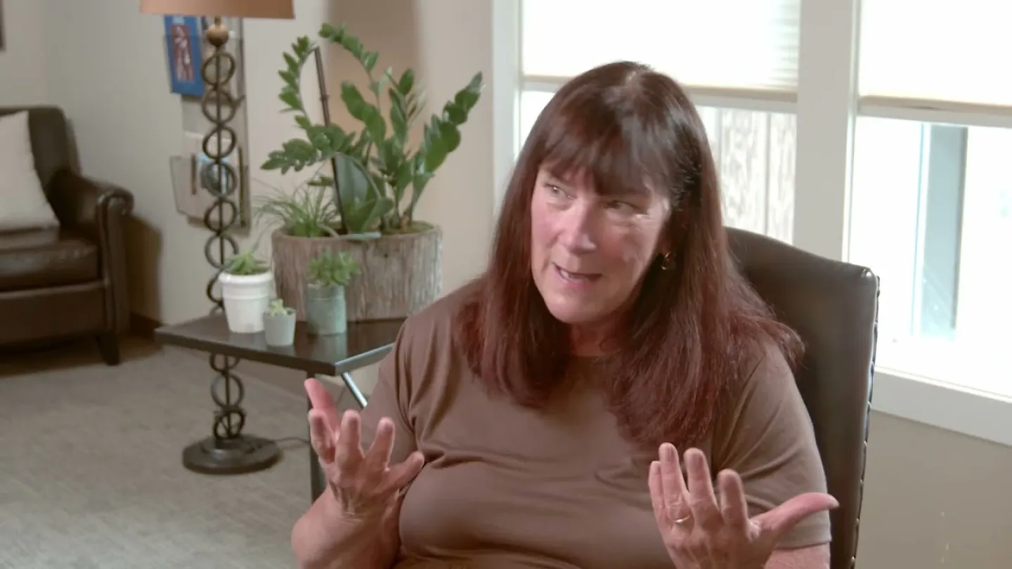 Woman with long brown hair talking animatedly while seated in a living room with plants and natural light.