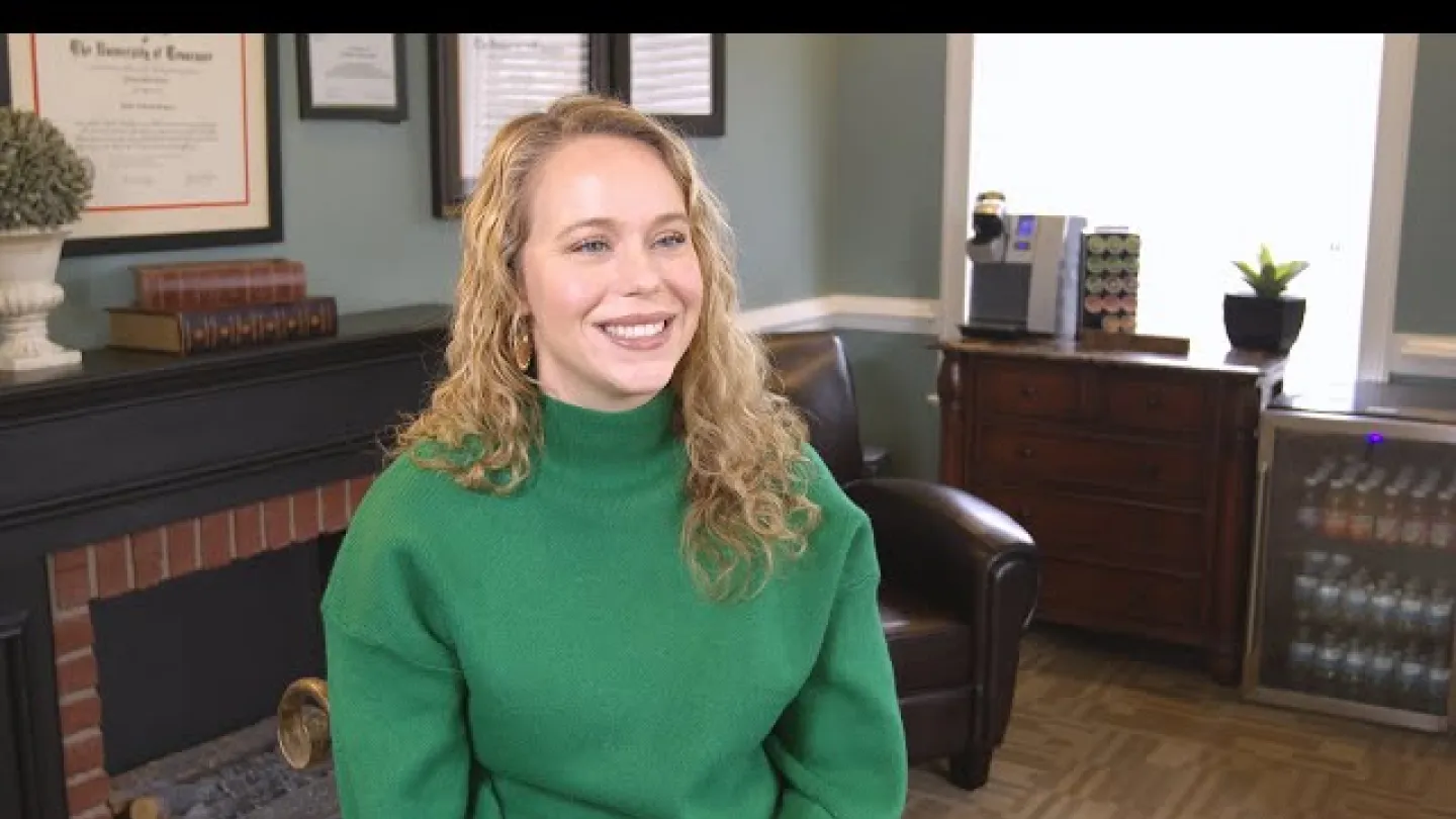 Smiling woman with curly blonde hair wearing a green sweater sitting in a cozy office room.