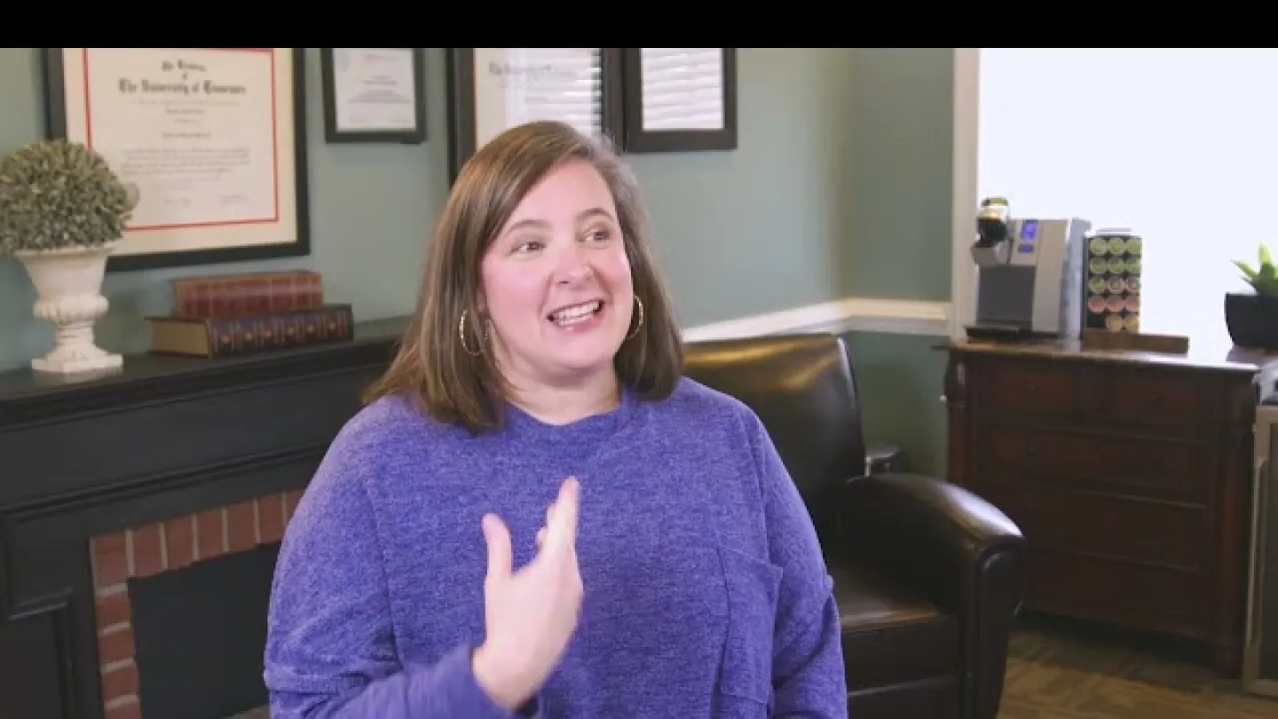Smiling woman in a blue sweater speaking in a cozy office with framed certificates and leather chair.