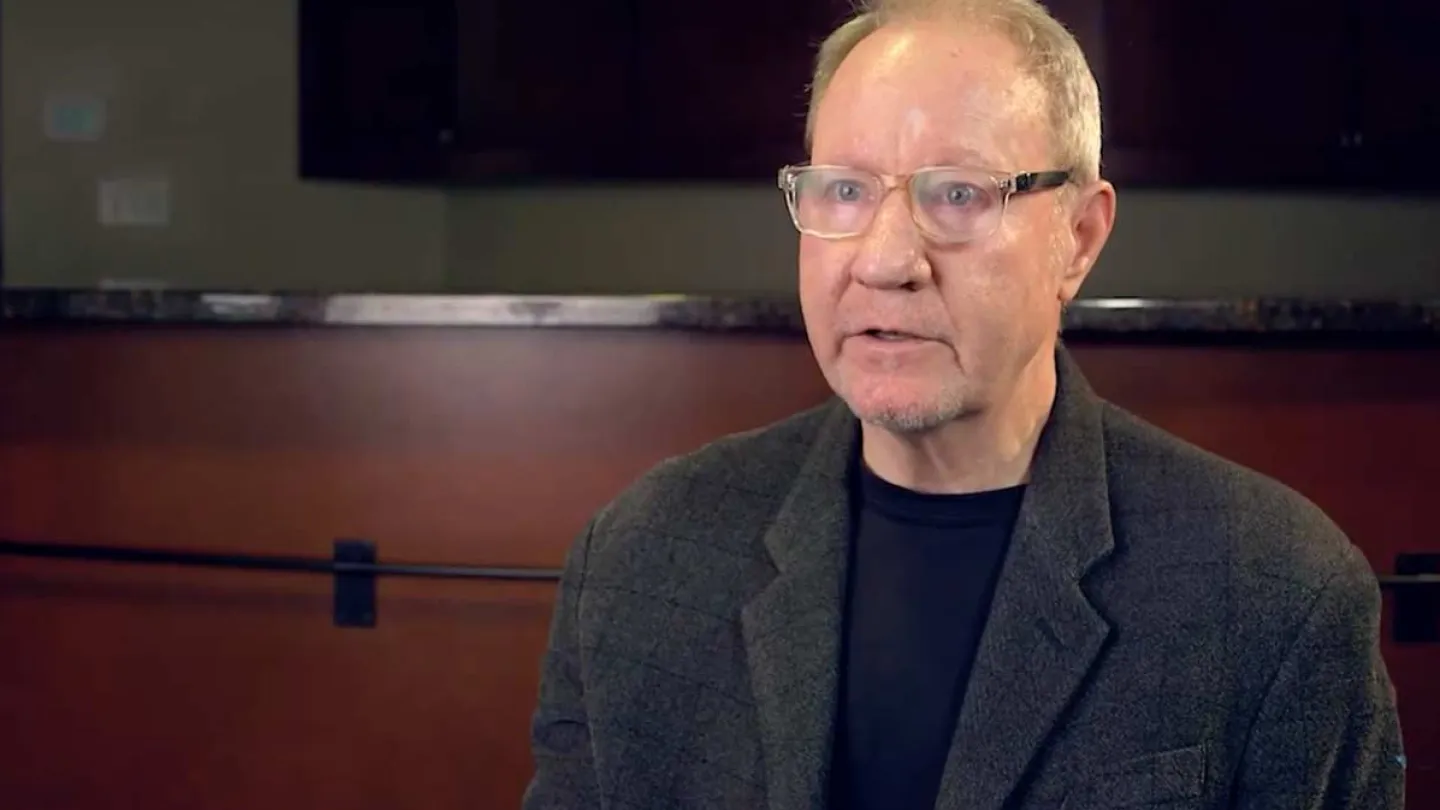 Middle-aged man with glasses wearing a dark blazer and shirt, speaking in an indoor setting with dim lighting.