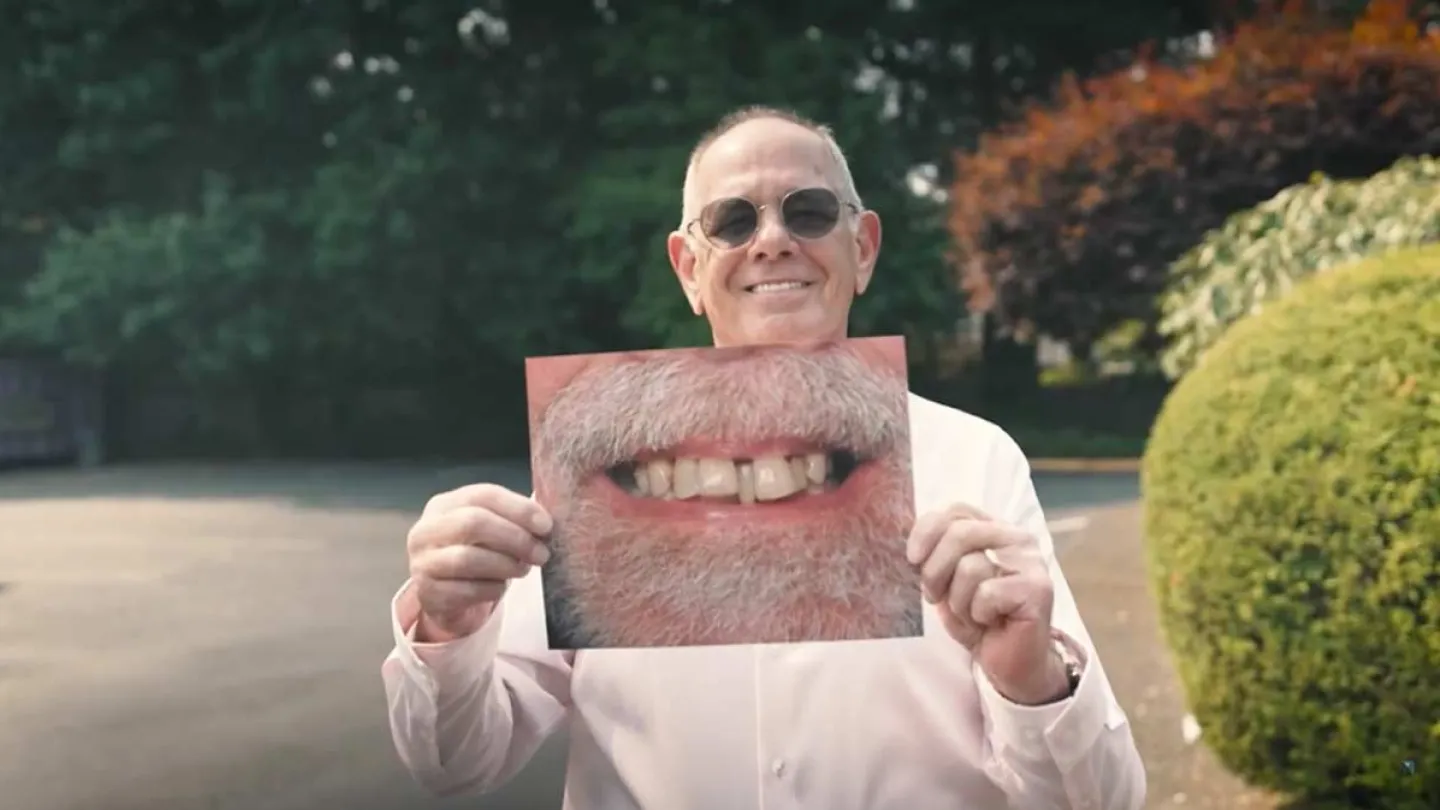 Smiling man wearing sunglasses holds photo of a bearded mouth with missing teeth outdoors in a park.