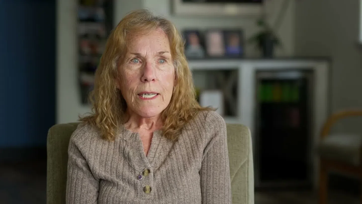 Older woman with curly blond hair wearing a taupe sweater sitting in an indoor living room setting.
