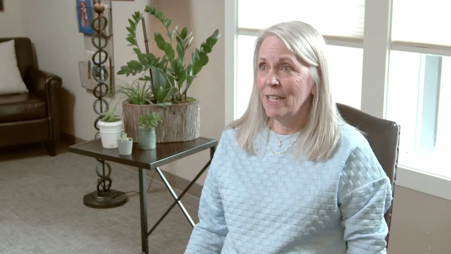 Smiling woman with gray hair sitting indoors near window and table with potted plants in a cozy living room.