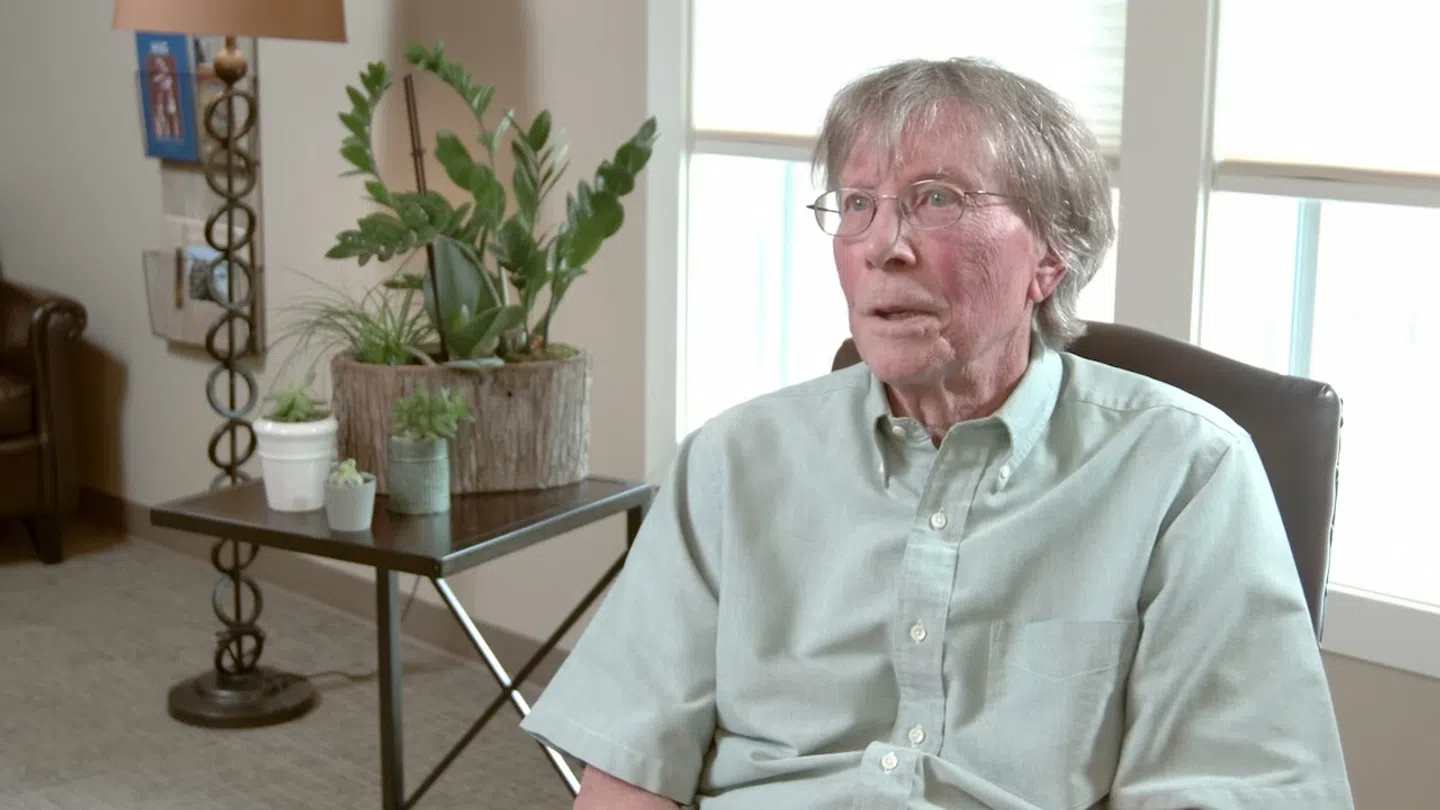 Elderly man with glasses sitting in a light-filled room next to a table with potted plants and a lamp.