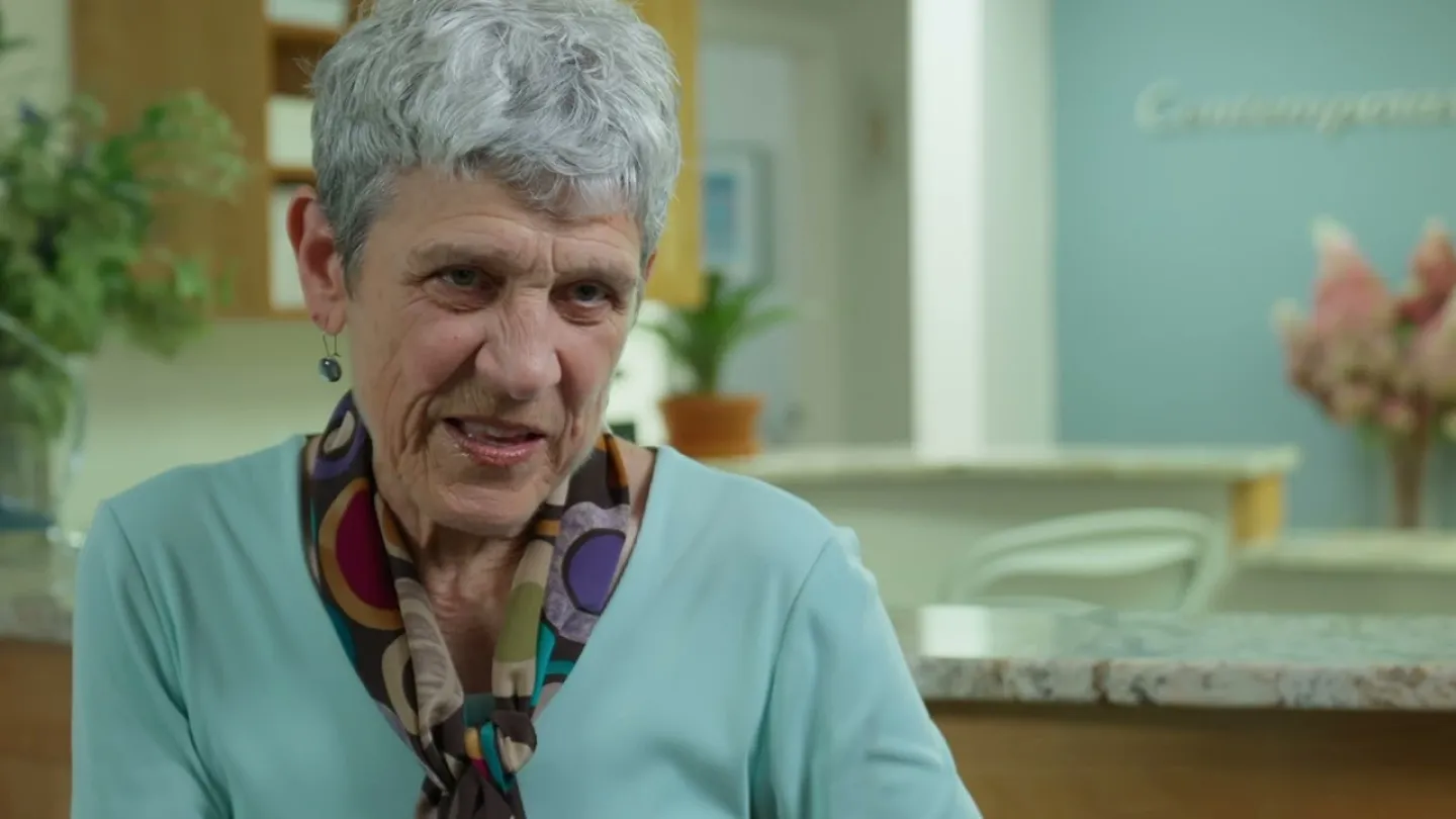 Elderly woman with gray hair wearing a colorful scarf and turquoise top in a bright, cozy room.