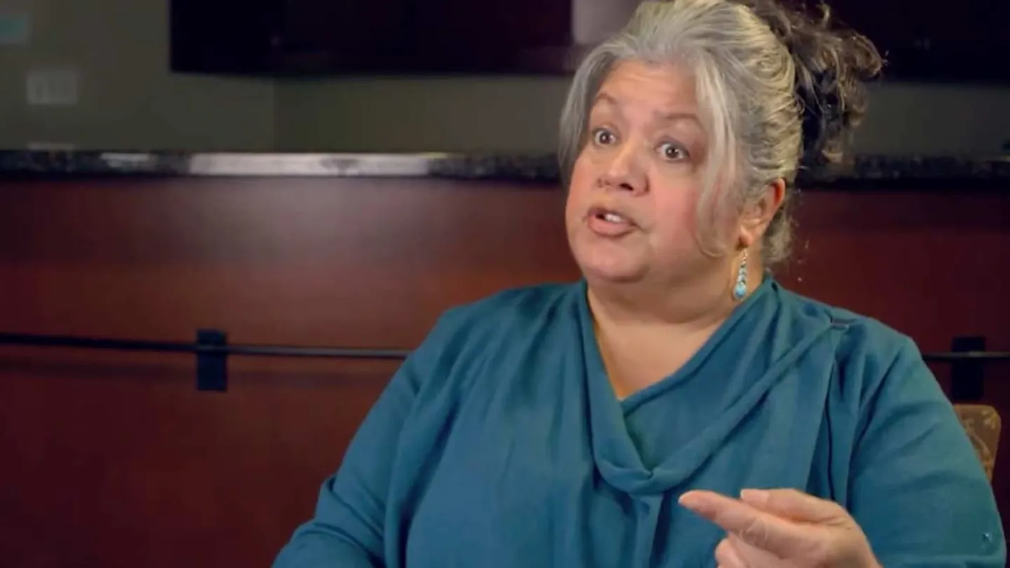 Middle-aged woman in teal top speaks expressively indoors, gesturing with her hand and wearing earrings.