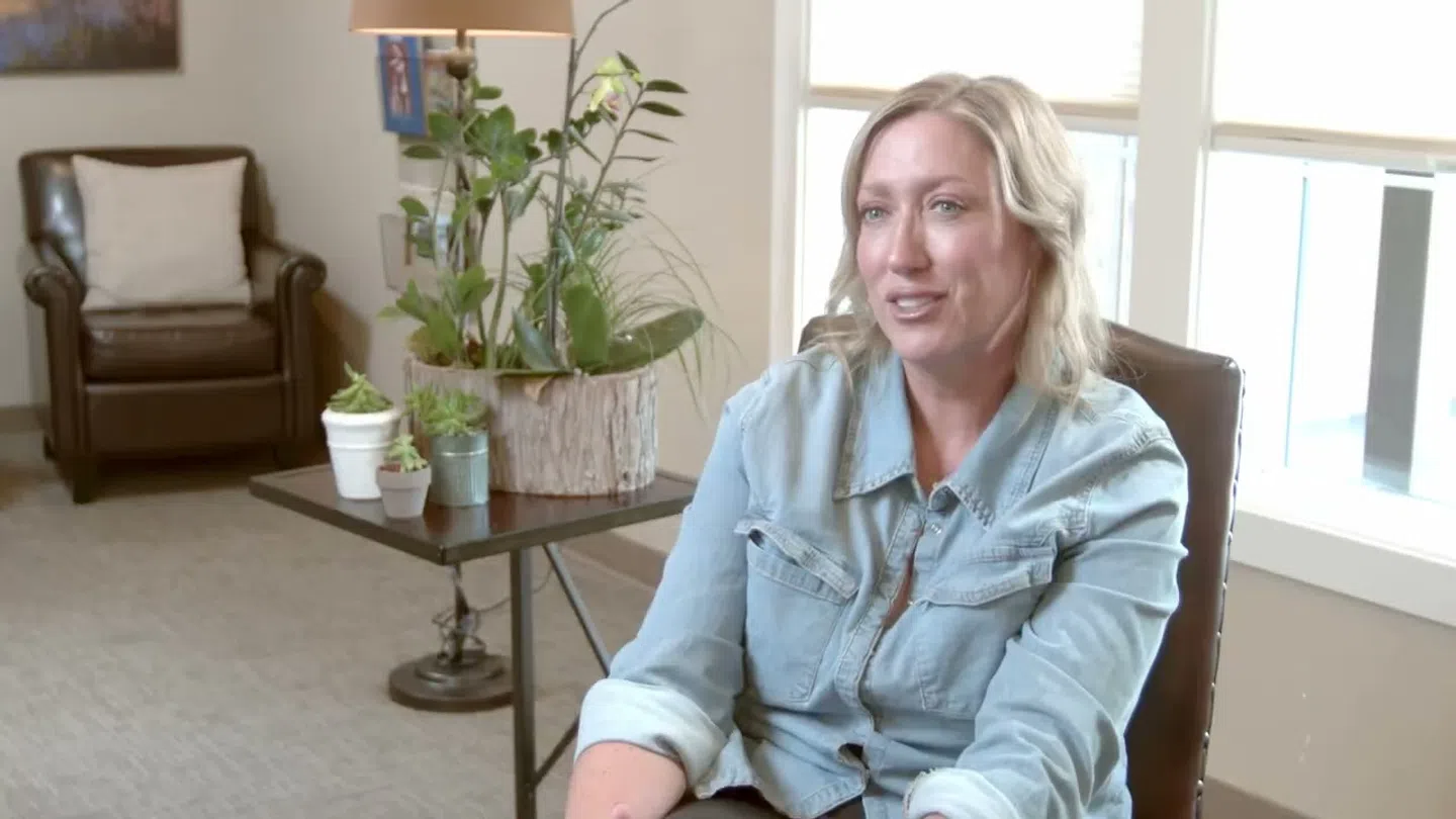Woman in a light denim shirt sitting and talking in a cozy, plant-filled living room with natural light.