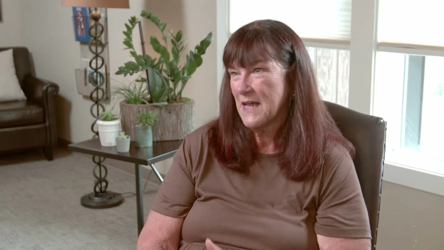 Middle-aged woman with brown hair talking while seated in a bright, cozy living room with plants and furniture.