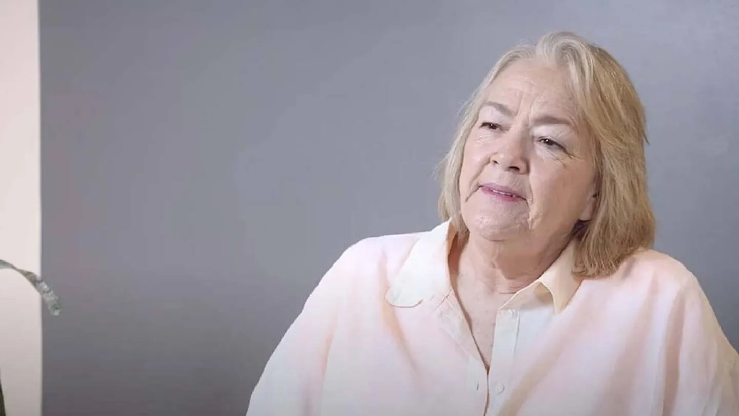 Older woman with blonde hair wearing a light pink shirt against a gray background looking thoughtfully.