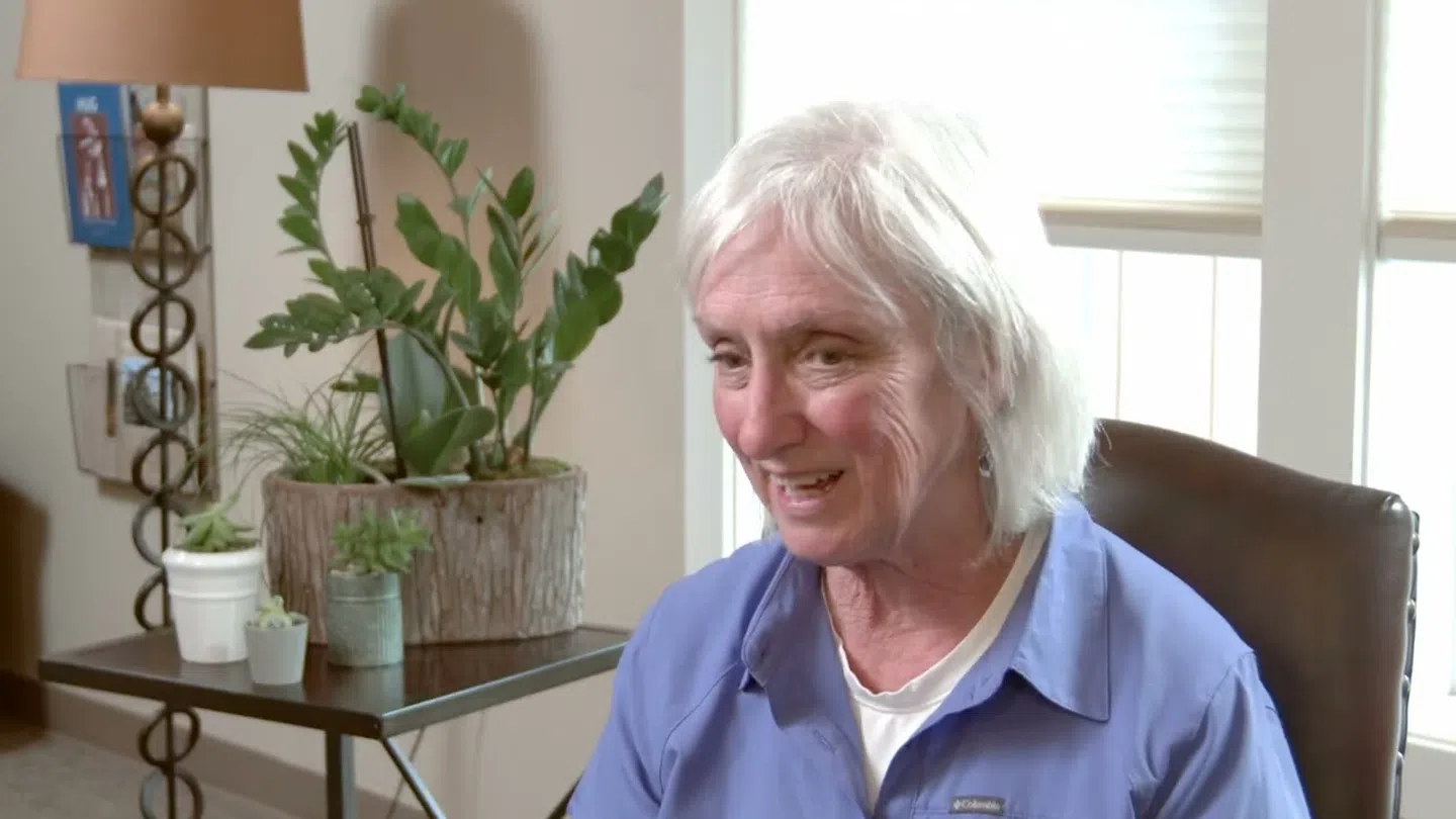 Elderly woman with white hair smiling, sitting in chair near window and indoor plants on table behind her.